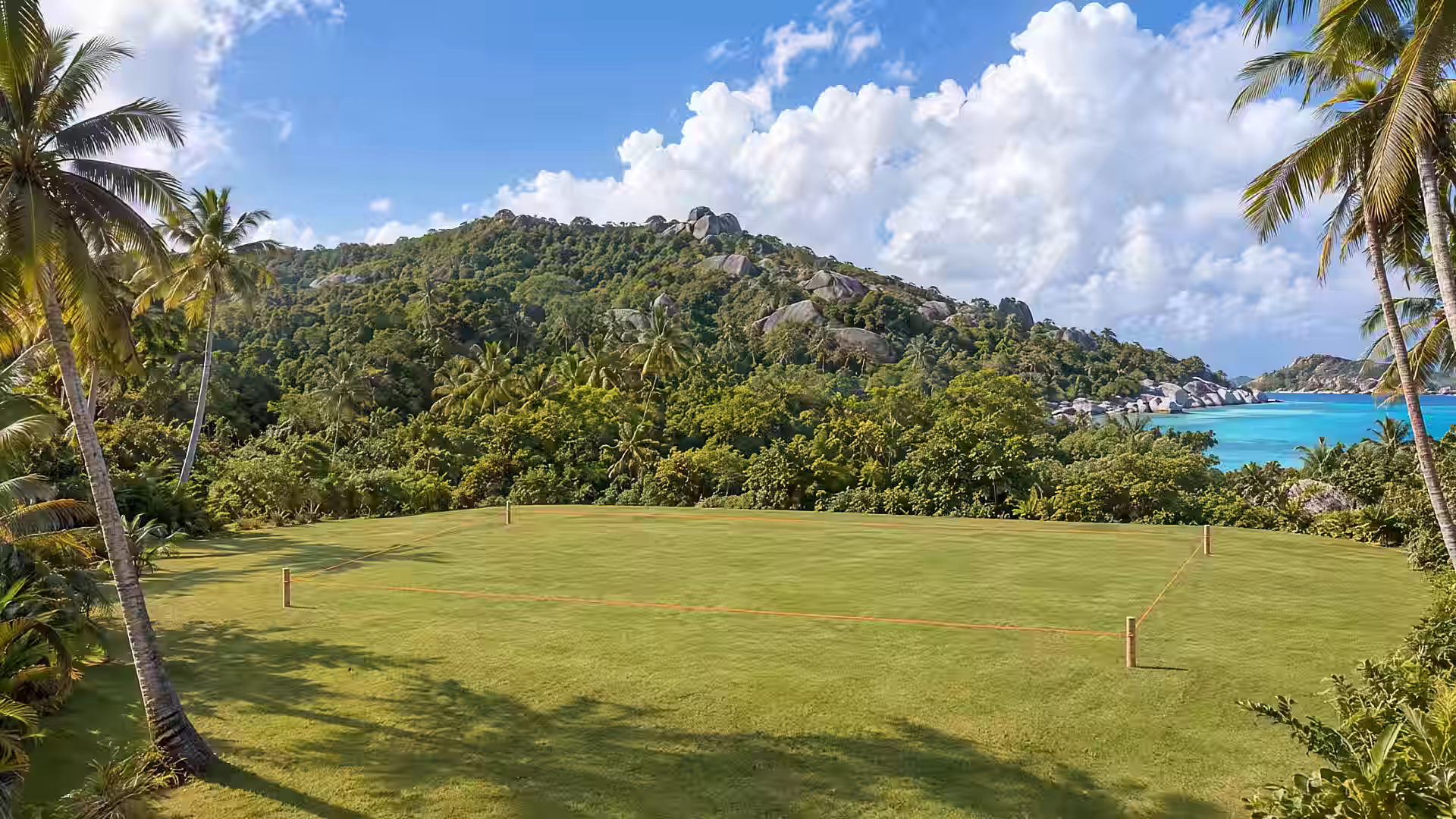 An empty, freshly mown plot of grass framed by coconut palms, with a forested granite hill and a turquoise lagoon behind. Wooden pegs and an orange string mark out where the house will stand.